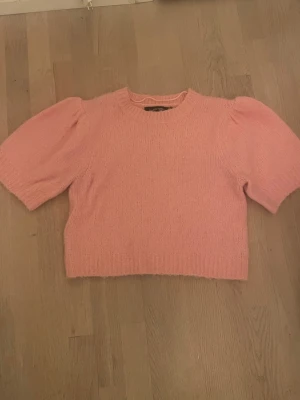 Rosa stickad tröja med puffärm - Superfin rosa stickad tröja från Vero Moda  med korta puffärmar och rund halsringning. Tröjan har en mjuk och fluffig känsla och ribbstickade detaljer vid ärmslut och nederkant. Perfekt för att addera färg till din outfit. Skulle säga att den är pyttelite nopprig men skulle inte säga att det syns (se bilder) Kan gå ner i pris!