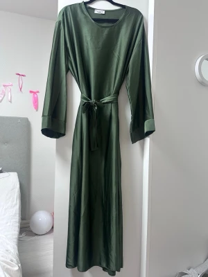Grön långklänning med knytband - Stilren långklänning i mörkgrönt satinliknande material med rund halsringning och vida långärmar. Knytband i midjan ger en snygg siluett. Perfekt för dig som gillar en elegant och enkel look.