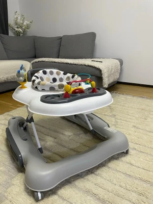 Gåstol med lekbräda och prickigt tyg - Säljer en gåstol i plast med grå och vita detaljer samt en lekbräda med färgglada leksaker. Sitsen är mjuk med svart, grå och beige prickigt tyg. Perfekt för små barn som vill utforska och leka.