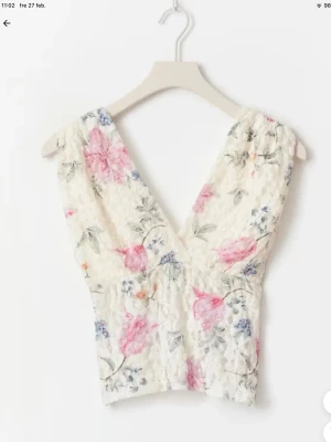 Blommig spets topp med v-ringning - Romantisk blommig topp med v-ringning och ärmlös design. Toppen är i vit spets med rosa, blå och gröna blommor och har en croppad passform. Perfekt för dig som gillar en feminin och somrig stil. Storlek m