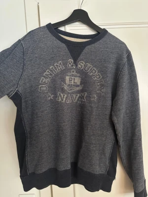 Grå sweatshirt Denim & Supply RL - Mörkgrå sweatshirt från Denim & Supply Ralph Lauren med tryckt marin-inspirerat motiv och text på bröstet. Tröjan har rund halsringning, ribbade muddar och kontrastdetaljer i svart vid sidorna och halsen. Perfekt för en avslappnad och mycket trendig stil. Storleken är L men den sitter mer som storleken M.