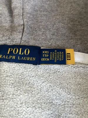 Grå zip hoodie från Polo Ralph Lauren - Snygg grå hoodie från Polo Ralph Lauren med dragkedja, huva och vita dragsnören. Klassisk blå logga broderad på bröstet och två fickor framtill.