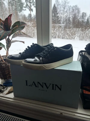 Svarta Lanvin sneakers med lackdetaljer - Snygga svarta sneakers från Lanvin med glansiga lackdetaljer på tån och klassisk vit sula. Skorna har lågt skaft, svarta skosnören och en stilren design som passar till det mesta. Materialmix av textil och lack ger en exklusiv känsla.