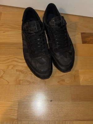 Svarta sneakers från Valentino - Snygga svarta sneakers från Valentino med kamouflagemönster och svarta skosnören. Skorna har en klassisk låg siluett och är tillverkade i en mix av mocka och textil. Perfekta för dig som gillar stilrena och exklusiva sneakers med en unik touch.