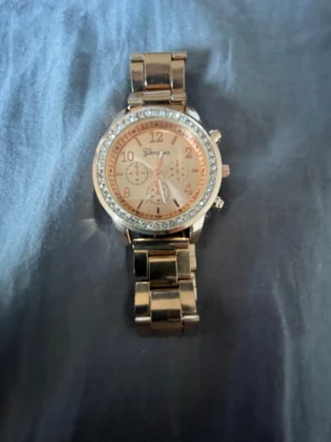 Roséguld klocka från Geneva - Snygg klocka från Geneva i roséguld med glittrande stenar runt urtavlan. Klockan har metallarmband och flera dekorativa urtavlor. Perfekt accessoar för att lyfta din stil.