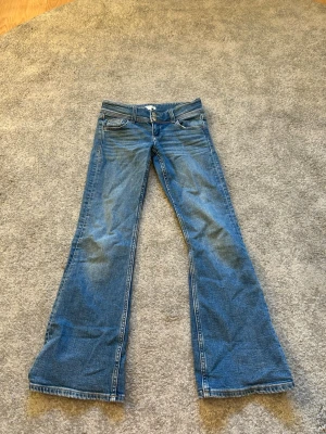 Blå bootcut jeans från Gina Tricot - Säljer ett par blå bootcut jeans från Gina Tricot med dubbla knappar i midjan och klassiska fickor fram och bak. Jeansen har en lätt utsvängd passform och är tillverkade i denim. Perfekta för en avslappnad och trendig look.