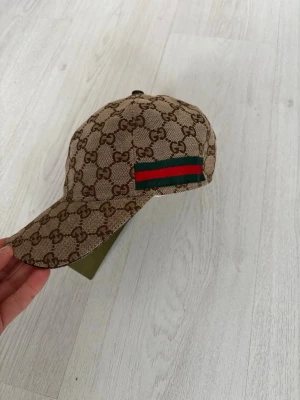 Beige Gucci Keps - Snygg beige keps från Gucci med enjusterbar rem bak och böjd skärm. Tillverkad i canvasmaterial med insida i ljus textil. Perfekt för till sommaren. OBS!!! Priset är rörligt