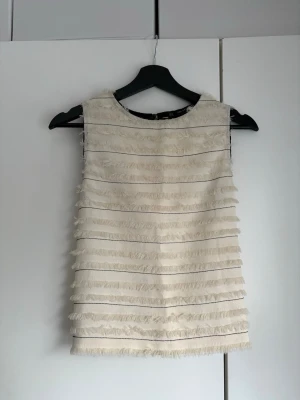 Beige ärmlös topp med fransar - Säljer en beige ärmlös topp med fransdetaljer över hela plagget. Toppen har rund halsringning och en rak, croppad passform. 