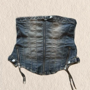 Jeanskorsett med dragkedjor - Cool jeanskorsett i blå denim med markerade sömmar och dubbla dragkedjor framtill. Justerbara remmar och tryckknappar i sidorna ger en edgy look. Perfekt för dig som vill ha en unik och trendig stil.