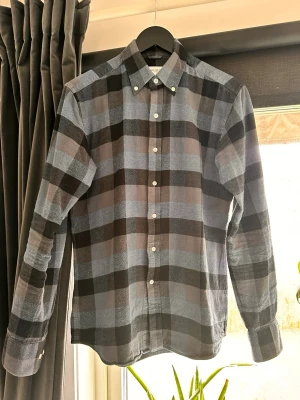 John Henric rutig skjorta - Snygg skjorta med stora rutor i blått, svart och grått. Klassisk krage med knappar och lång ärm. Perfekt för dig som gillar en avslappnad men ändå stilren look. Skjortan har knäppning framtill och är tillverkad i mjukt bomullstyg.