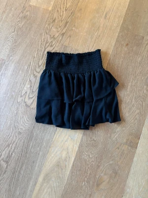 Svart volangkjol med smock - Kort svart kjol med bred smock i midjan och dubbla volanglager. Kjolen har en luftig och flowy känsla, perfekt för att styla med crop tops eller stickade tröjor. Materialet känns lätt och bekvämt, och den elastiska midjan gör den enkel att bära.