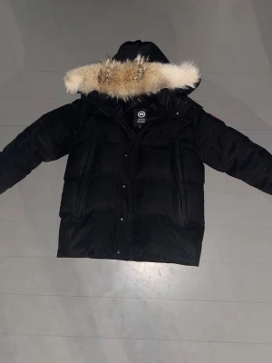 Canada Goose ”WYNDHAM” 1:1 A - Säljer en svart dunjacka från Canada Goose med stor huva och fluffig pälsdetalj. Jackan har flera fickor framtill och klassisk Canada Goose-logga på ärmen. Perfekt för kalla vinterdagar och riktigt varm tack vare dunfyllningen, ända negativa e dragkedjan, den e lite trög men de är knappt ett problem. Scan finns, fin päls som inte känns eller ser fejk ut, kom pm för mer bilder och om ni vill se scan-taggen! OBS Pris diskuteras