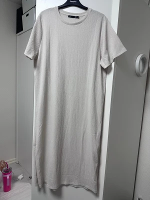 Ljusbeige maxi klänning/tshirtklänning - En stilren, ljusbeige klänning i maxi modell. Klänningen har korta ärmar, rund halsringning och är tillverkad i ett strukturerat, mjukt material. Perfekt för en avslappnad och enkel look.
