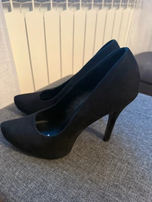 Svarta klassiska pumps med klack - Säljer ett par stilrena svarta pumps med spetsig tå och hög smal klack. Skorna har en elegant siluett och är tillverkade i ett material som ser ut som mocka. Perfekta för dig som gillar en tidlös look.
