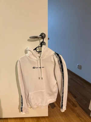 Vit hoodie från Champion med logga - Snygg vit hoodie från Champion med svart logga broderad på bröstet och svarta band med Champion-tryck längs ärmarna. Klassisk känguruficka framtill och justerbar huva med snören. Perfekt för en chill och sportig look.