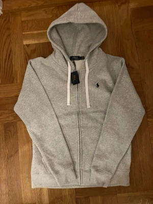 Polo Ralph Lauren zip hoodie - Snygg grå hoodie från Polo Ralph Lauren med klassisk broderad logga på bröstet. Tröjan har huva med vita snören, hel dragkedja och ribbade muddar. Perfekt för chill dagar och enkel att matcha med jeans eller joggers.