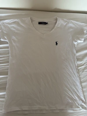  Polo Ralph Lauren - Klassisk vit t-shirt från Polo Ralph Lauren med v-ringning och korta ärmar. T-shirten har den ikoniska broderade loggan i marinblått på bröstet. Tillverkad i mjuk bomull för en skön känsla och enkel stil. Den är använd en gång, jag köpte den i somras och jag är själv storlek xs-s men den passar bra som M för den är väldigt tajt