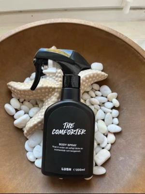 Lush The Comforter Body Spray 🫐🍦200 ml - The Comforter Body Spray från Lush i en svart plastflaska med sprayfunktion. Doften har inslag av svartvinbär och bergamott. Flaskan rymmer 200 ml och har en modern, stilren design med vit text på framsidan. Luktar gourmandig mjuk blåbärspaj och godisaktig! Ca 70% kvar. 