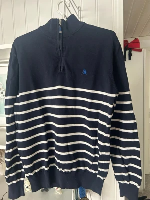 Marinblå randig tröja med halvzip - Snygg marinblå tröja från Sail Racing med vita horisontella ränder och halvzip framtill. Tröjan har en liten blå logga med segelbåt på bröstet, ribbade muddar och hög krage. Perfekt för en avslappnad men stilren look.