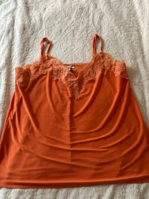 Orange linnetopp med spetsdetaljer - Snygg orange linnetopp med tunna axelband och v-ringad spetskant framtill och baktill. Toppen har en mjuk och följsam känsla, perfekt för varma dagar. Spetsen ger en extra touch av stil och gör toppen lätt att matcha med olika outfits.