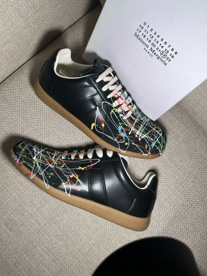 Maison Margiela svarta sneakers med färgstänk - Maison Margiela sneakers i svart läder med vita snören och coola färgstänk i rött, gult, blått och vitt. Klassisk låg modell med beige gummisula och vit insida. Perfekt för dig som vill sticka ut med en unik och trendig look.