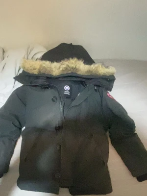 Svart parkas från Canada Goose - Säljer en svart parkas från Canada Goose med huva och fuskpälsdetalj. Jackan har dragkedja och knappar framtill samt en klassisk patch på ärmen. Perfekt för kalla dagar med sin tjocka vaddering och tidlösa design.