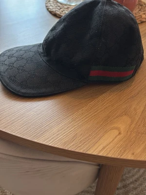 Svart Gucci keps med GG-mönster - Säljer en svart keps från Gucci med klassiskt GG-monogram över hela kepsen. På sidan finns den ikoniska gröna och röda randen. Kepsen har böjd skärm och är tillverkad i ett slitstarkt tygmaterial. Perfekt för dig som vill ha en stilren och lyxig accessoar.