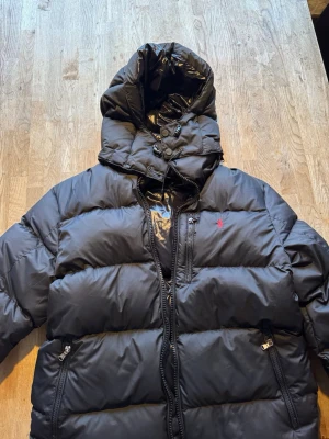 Svart dunjacka från Polo Ralph Lauren - Säljer en svart dunjacka från Polo Ralph Lauren med huva och dragkedja framtill. Jackan har två sidofickor med dragkedja och den klassiska röda loggan på bröstet. Puffig modell som håller dig varm under kalla dagar.