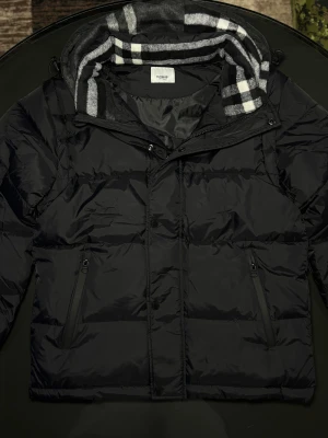 Svart pufferjacka från Burberry - Svart pufferväst från Burberry med avtagbar huva i klassiskt rutigt mönster. Västen har dragkedjor på fickorna och knappar framtill. Materialet är polyester och fodret är mjukt och skönt. Perfekt för dig som vill ha en clean och stilren look.