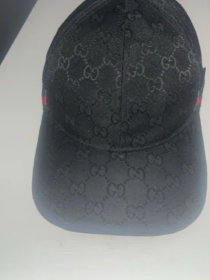 Svart Gucci keps med GG-mönster - Snygg svart keps från Gucci med det klassiska GG-monogrammet över hela kepsen. Kepsen har böjd skärm, justerbart spänne bak och diskreta röda och gröna detaljer på sidorna. Tillverkad i ett slitstarkt tyg som ger en exklusiv känsla.
