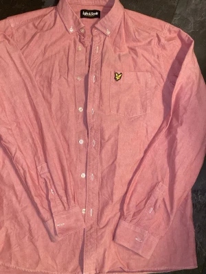 Rosa skjorta från Lyle & Scott L - Säljer en rosa skjorta från Lyle & Scott med klassisk button-down krage och bröstficka med den ikoniska gula örnloggan. Skjortan har långa ärmar och vita knappar. Perfekt för en stilren look.