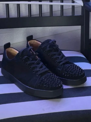 Christian Louboutin skor  - Säljer ett par svarta sneakers med coola svarta nitar på tån. Skorna har snörning, platt sula och är gjorda i mockaliknande material. Insidan är beige och de har en modern, edgy look som sticker ut.