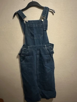 Mörkblå jeansbyxdress med hängslen - Snygg mörkblå jeansbyxdress med klassiska hängslen och bröstficka. Byxdressen har raka ben, sidofickor och justerbara axelband med metallspännen. Perfekt för en avslappnad och trendig look.