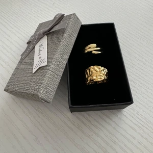 Guldiga ringar i presentask - Två stilrena guldiga ringar i en elegant presentask. Den ena ringen har en slät, modern design medan den andra har en texturerad yta som ger en unik look. Perfekt som present eller för att lyxa till din egen smyckessamling!