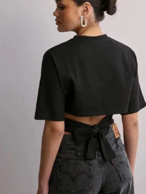Svart croppad topp med knyt från Nelly - Cool svart croppad topp från Nelly med korta ärmar och snygg knytning i ryggen. Perfekt för dig som gillar trendiga och stilrena plagg. Materialet är mjukt och känns bekvämt mot huden.