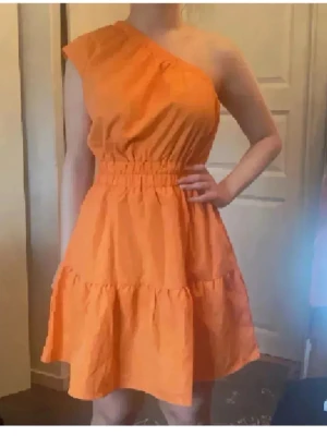 Finaste sommarklänningen❤️ - Säljer en snygg orange kortklänning med oneshoulder-design och resår i midjan. Klänningen har volangdetaljer på kjolen och är tillverkad i ett lätt tyg som passar perfekt till varma dagar. Enkel men trendig modell som sticker ut. Köpt utomlands