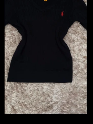 Marinblå stickad tröja från Ralph Lauren - Klassisk marinblå stickad tröja från Ralph Lauren med röd broderad logga på bröstet. Tröjan har v-ringning och ribbstickade muddar vid comb och nederkant. Perfekt för en stilren och tidlös look.