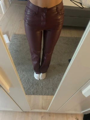 Vinröda skinnbyxor från BinBok - Snygga vinröda skinnbyxor med hög midja. Byxorna har klassisk femficksdesign och stängs med knapp och dragkedja. Perfekta för dig som vill ha en trendig och edgy look.