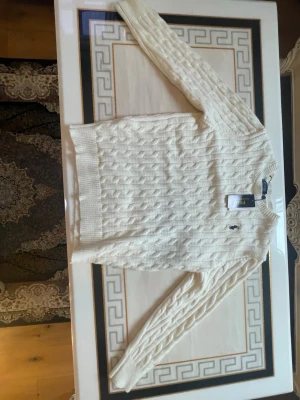 Vit kabelstickad tröja Polo Ralph Lauren - Kabelstickad vit tröja från Polo Ralph Lauren med klassisk rund halsringning och broderad logga på bröstet. Långa ärmar och ribbade muddar vid ärmslut och nederkant. Perfekt för lager på lager och stilrena outfits.