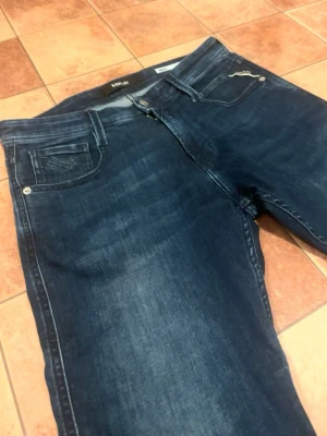 Replay Anbass - Snygga mörkblå jeans från Replay med klassisk femficksdesign och diskreta slitningar. Jeansen har raka ben och detaljerade sömmar, samt en läderpatch med Replay-logga bak i midjan. Perfekta för en trendig och avslappnad look.