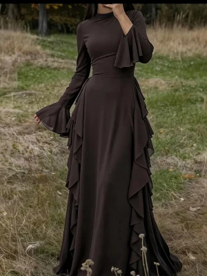Mörkbrun långklänning med volanger - oanvänd - Elegant långklänning i mörkbrunt med långa trumpetärmar och volangdetaljer längs sidorna. Klänningen har en figurnära passform upptill och faller mjukt från midjan. Perfekt för dig som vill sticka ut med en unik och stilren look.