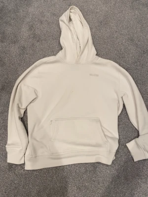 Vit hoodie från Hollister - Snygg vit hoodie från Hollister med diskret logga på bröstet. Hoodien har en stor känguruficka med dragkedja på ena sidan och klassisk huva. Tillverkad i mjukt bomullsmaterial som känns skönt mot huden. Perfekt för en avslappnad och stilren look.
