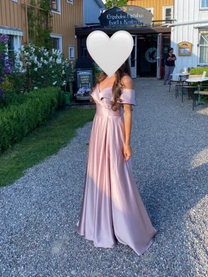 Ljuslila axelbandslös långklänning - Elegant ljuslila långklänning i satin med offshoulder och v-ringad urringning. Klänningen har en figurnära överdel och ett utsvängt, svepande kjolparti som ger en lyxig känsla. Perfekt för bal eller student.