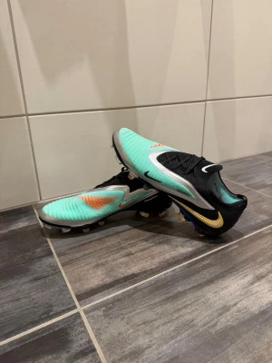 Nike Phantom GT2 fotbollsskor turkos - Säljer ett par Nike Phantom GT2 fotbollsskor i en snygg turkos och svart färg med orange och guldiga detaljer. Skorna har en låg profil, snörning och är tillverkade i syntetmaterial för bra bollkontroll. Perfekta för dig som vill sticka ut på planen.