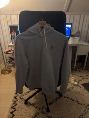 Grå hoodie från Polo Ralph Lauren - Snygg grå hoodie från Polo Ralph Lauren med klassisk huva och dragsnören. Har en liten blå logga broderad på bröstet och en stor magficka framtill. Perfekt för chill dagar och enkel att matcha med jeans eller joggers.