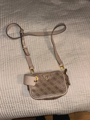 Beige monogram crossbody från Guess - Snygg beige crossbody-väska från Guess med klassiskt monogrammönster och guldfärgade detaljer. Väskan har justerbar axelrem i matchande färg och Guess-logga i metall framtill. Tillverkad i konstläder med struktur och dragkedja upptill.