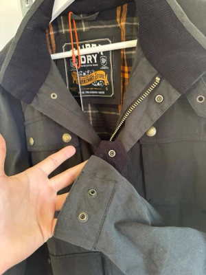 Grå jacka från Superdry Merchant Store - Snygg grå jacka från Superdry Merchant Store med rutigt innerfoder i orange, grått och vitt. Jackan har tryckknappar, dragkedja och detaljer i manchester på kragen. Perfekt för dig som gillar klassisk stil med moderna detaljer. Jackan är köpt i london därav har jag inte kunnat lämna tillbaka den, aldrig använt den så helt ny! Köpt för 1699kr