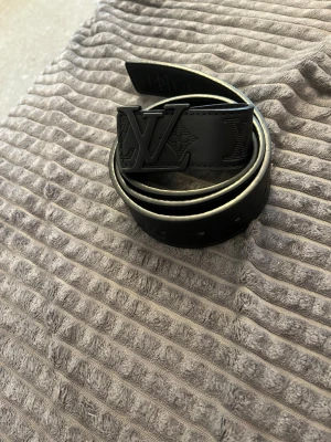 Svart Louis Vuitton skärp i skinn - Snyggt svart skärp från Louis Vuitton med präglad logga och monogramdetaljer längs hela bandet. Skärpet har ett stort LV-spänne i svart och är gjort i skinn. Perfekt accessoar för att lyfta vilken outfit som helst.
