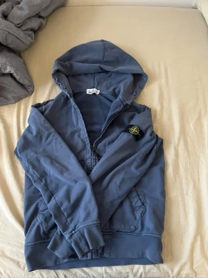 Blå hoodie från Stone Island - Snygg blå hoodie från Stone Island med dragkedja och klassisk logotyp-patch på vänster ärm. Hoodien har huva, känguruficka och ribbade muddar. Perfekt för en avslappnad och trendig stil. Materialet känns mjukt och bekvämt mot huden.