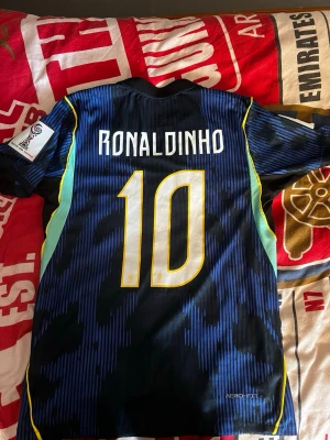 Blå Brasilien fotbollströja Ronaldinho - Snygg blå Brasilien fotbollströja från Nike med Ronaldinho och nummer 10 på ryggen. Tröjan har gula detaljer, CBF-logga och Aerofit-material för extra komfort. Perfekt för dig som älskar fotboll och vill sticka ut på planen.